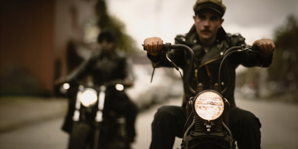 Two riders on bobbers rolling toward camera, front headlights lit, Brake House leather jackets