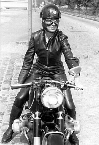Person in leather outfit and helmet sitting on a motorcycle on a road.