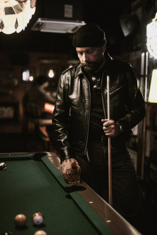 Rider in a leather jacket and beanie with a whiskey glass and cue at the pool table