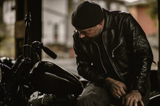 Rider in a beanie and black leather jacket leaning over the tank in a garage