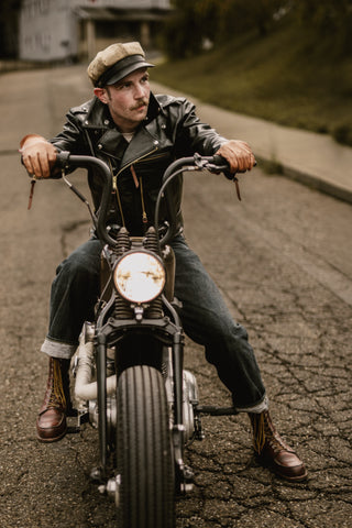 Rider in a black leather jacket and cap on his bobber, brown leather boots on the pegs