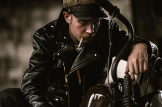 Rider seated on his bobber with a cigarette, black leather jacket and cap
