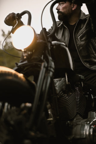 Bearded rider in a black leather jacket beside a glowing headlight at sunset