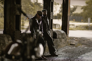 Two riders resting against a bridge pillar beside their bikes